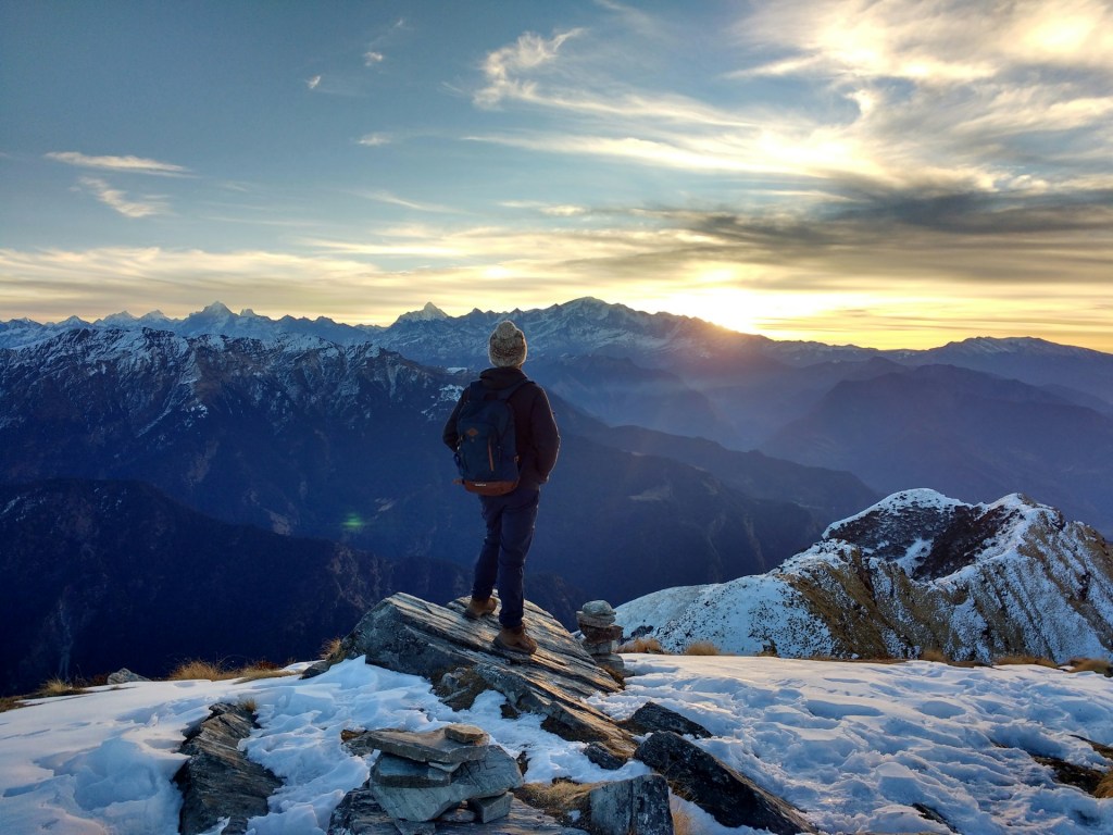 A person standing on a rocky outcrop overlooking a mountainous landscape at sunset, with snow covering some areas and distant peaks visible in the background.